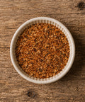 A top-down view of a small white ceramic bowl filled with a coarse seasoning blend, on a wooden table.