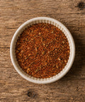 A top-down view of a small white ceramic bowl filled with a reddish-brown spice blend, on a wooden able.
