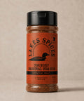 A 5oz bottle of Lake Spices Foundry Original BBQ Rub, featuring the company logo of a black loon on an orange background, and a black icon of Minnesota in the upper righthand corner. Label text above the loon reads “Lake Spices: Honest Blends for Homemade Taste.”