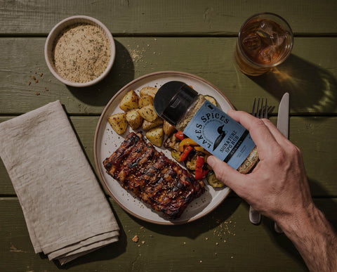 A hand sprinkling a bottle of Lake Spices Signature SPG Rub over a plate of grilled meat, roasted potatoes, and mixed vegetables on a dark green wooden table. A folded napkin, a bowl of seasoning mix, and an iced drink are visible nearby.
