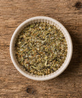 A top-down view of a white ceramic bowl filled with an herb and spice seasoning blend, on a wooden table.