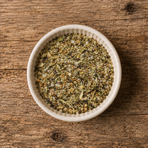 A top-down view of a white ceramic bowl filled with an herb and spice seasoning blend, on a wooden table.