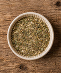 A top-down view of a white ceramic bowl filled with a greenish herb seasoning blend, on a wooden table