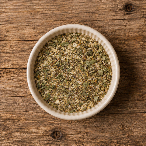 A top-down view of a white ceramic bowl filled with a greenish herb seasoning blend, on a wooden table