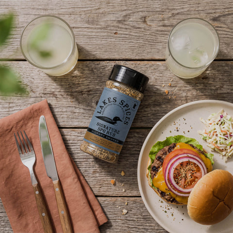 A top-down shot of a bottle of Lake Spices Signature SPG Rub, a plated burger, and iced drinks, on a wooden table.