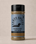 A 6oz bottle of Lake Spices Hickory Smoked Signature SPG Rub, featuring the company logo of a black loon on a blue-grey background, and a black icon of Minnesota in the upper righthand corner. Label text above the loon reads “Lake Spices: Honest Blends for Homemade Taste.”