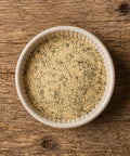 A top-down view of a light beige seasoning blend with visible green herbs, served in a small white ceramic bowl on a rustic wooden surface.