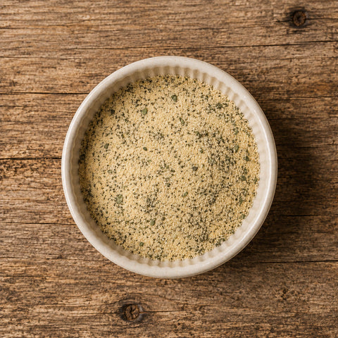 A top-down view of a light beige seasoning blend with visible green herbs, served in a small white ceramic bowl on a rustic wooden surface.