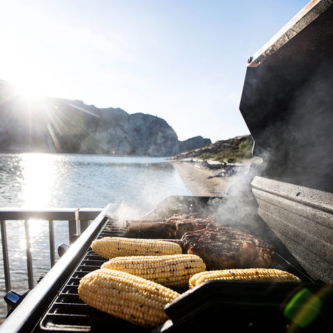An outdoor shot of a black charcoal grill with meat and corn on the cob cooking on the grate, against a sunny coastline background.