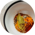 A close-up circular view of a white bowl containing a mixture of ground spices, including bright orange and red powders, mixed with coarse salt and a bay leaf.
