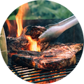 An outdoor shot of a black charcoal grill with meat cooking on the grate, with visible flames, and a hand holding tongs in the foreground.
