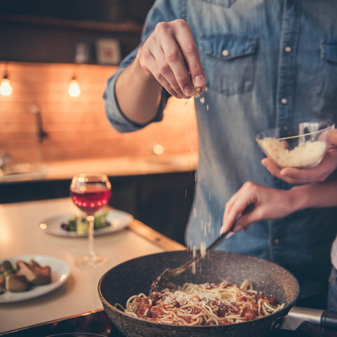 A close-up lifestyle shot of a person in a blue shirt, seasoning a pan of spaghetti from a small glass bowl of grated cheese.