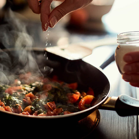 A close-up lifestyle shot of a person in a kitchen, seasoning a pan of cooked vegetables from a small glass jar of salt.