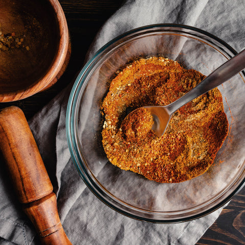 A top-down view of a small glass bowl filled with a reddish-brown seasoning blend, on a grey linen cloth next to a wooden mortar and pestle.