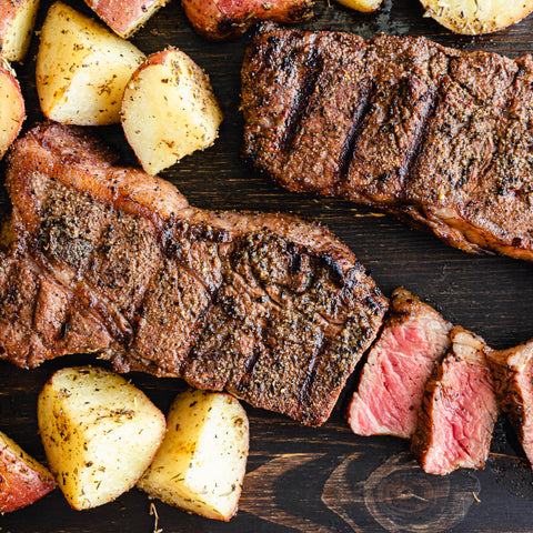 Two grilled steaks sliced and arranged on a dark wooden surface with potatoes.